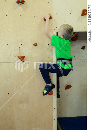 Rock climbing for children. Blond boy in a green shirt is climbing climbing wall. Bouldering class. Artificial track for safe activities and competitions. Mountaineering, safety system, special shoes 111601176