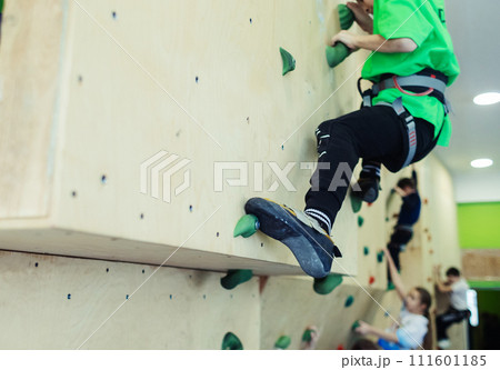 Rock climbing for children. Blond boy in a green shirt is climbing climbing wall. Bouldering class. Artificial track for safe activities and competitions. Mountaineering, safety system, special shoes Rock climbing for children. Blond boy in a green shirt is climbing climbing wall. Bouldering class. Artificial track for safe activities and competitions. Mountaineering, safety system, special shoes 111601185