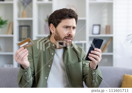 Bearded mature man appearing worried while holding credit card and smartphone, potentially facing financial issues. Bearded mature man appearing worried while holding credit card and smartphone, potentially facing financial issues. 111601711