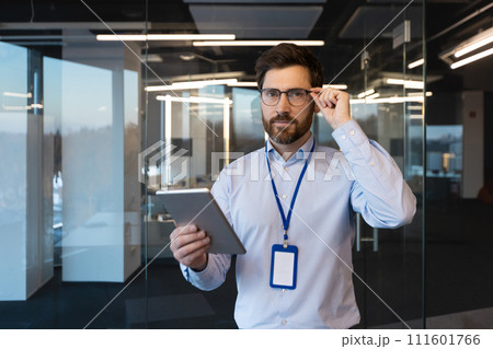 Focused mature businessman at his workplace in a modern office, adjusting his glasses while holding a digital tablet. 111601766