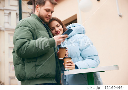 Beautiful lovely couple drinking coffee on terrace and man scrolling phone while looking for delivery service or shops online. 111604464