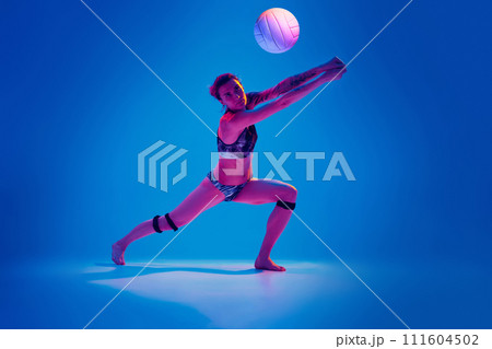Concentrated athlete woman, beach volleyball player passes ball from below against gradient blue background in pink neon light. 111604502
