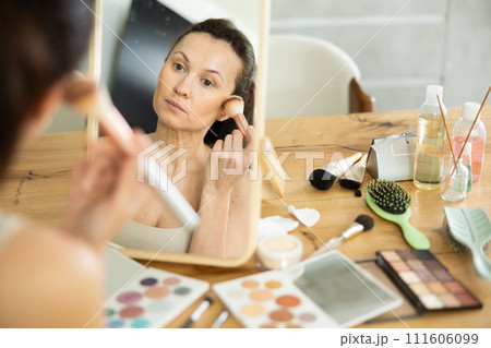 Middle-aged woman applying blush on her cheeks by means of makeup brush Middle-aged woman applying blush on her cheeks by means of makeup brush 111606099