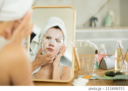 Middle-aged woman applying tissue mask on her facial skin sitting in front of the mirror 111606551
