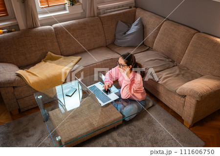 Top view of young Caucasian woman wearing glasses is sitting near sofa and using laptop. Concept of cozy remote work 111606750