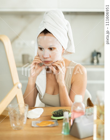 Middle-aged woman applying tissue mask on her facial skin sitting in front of the mirror 111606951