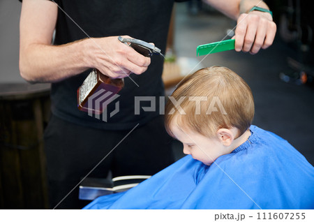 Male hairdresser sprinkling water on client's head. Barber using sprayer bottle with water to get hair wet and straight. Side view of little boy closing eyes to avoid water. Male hairdresser sprinkling water on client's head. Barber using sprayer bottle with water to get hair wet and straight. Side view of little boy closing eyes to avoid water. 111607255