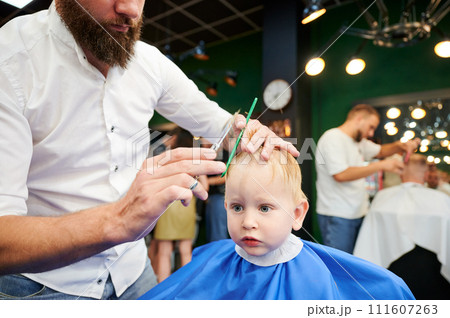 Man barber cutting little boy's hair using comb and scissors. Child getting haircut from adult male, likely barber. Professional hairdresser and cute client at modern barbershop. Man barber cutting little boy's hair using comb and scissors. Child getting haircut from adult male, likely barber. Professional hairdresser and cute client at modern barbershop. 111607263