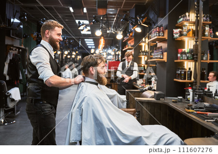 Regular client visiting favorite barbershop. Professional hairdresser and customer smiling. Male barber putting on white cape. 111607276