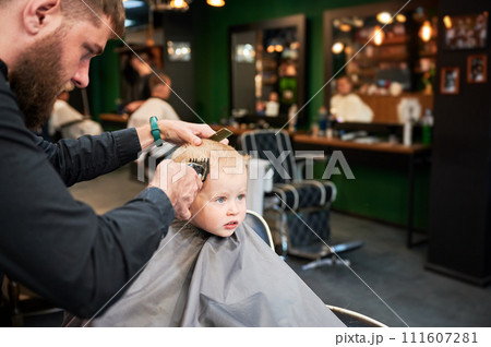 Barber using comb and shaver to cut hair. Professional hairdresser shaving little kid's nape. Barber using comb and shaver to cut hair. Professional hairdresser shaving little kid's nape. 111607281