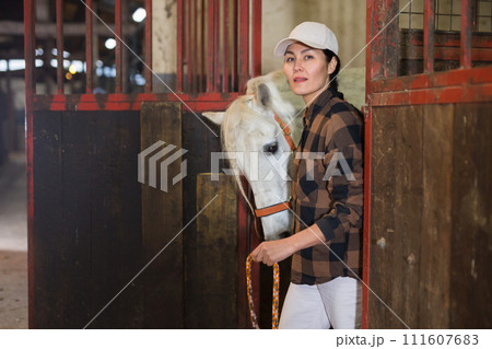 Horse breeder leading white horse through horse barn 111607683