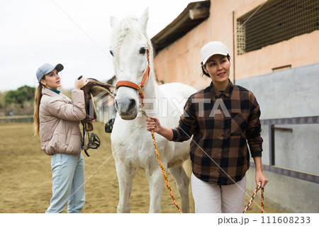 Asian female stable owner holding racehorse by bridle outdoor while girl putting saddle 111608233