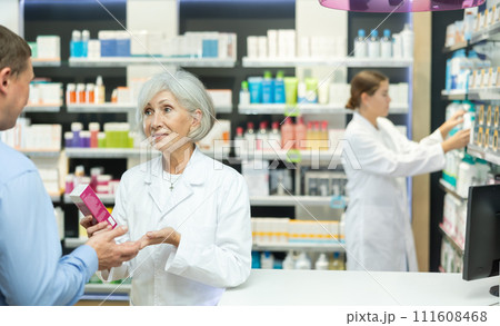 Mature female pharmacist consulting middle-aged man costumer about care product in box 111608468