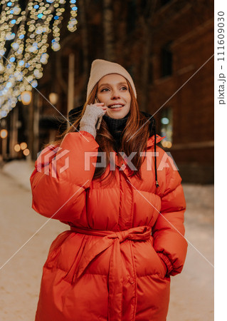 Vertical portrait of charming young woman in hat and winter jacket talking using mobile phone standing on dark snow city street on blurred background of hinged festive illumination, looking away. Vertical portrait of charming young woman in hat and winter jacket talking using mobile phone standing on dark snow city street on blurred background of hinged festive illumination, looking away. 111609030