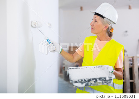 Portrait of elderly positive woman painter in protective helmet and yellow vest who paints walls with paint roller Portrait of elderly positive woman painter in protective helmet and yellow vest who paints walls with paint roller 111609031