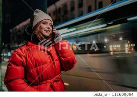Portrait of stylish young woman wearing hat and winter jacket using smartphone standing by road, on winter street of city at night. Beautiful redhead female calling taxi on blurred urban background. Portrait of stylish young woman wearing hat and winter jacket using smartphone standing by road, on winter street of city at night. Beautiful redhead female calling taxi on blurred urban background. 111609064