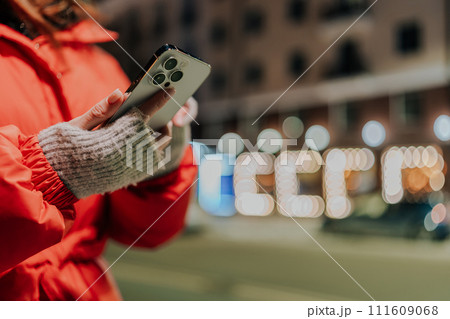 Closeup hands of unrecognizable woman typing on mobile phone at dark night city street on blurred background on bright bokeh light. Female in warm clothes typing text message in winter city nightlife. 111609068