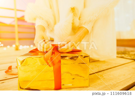 A woman in a white dress is holding a gold box with a red ribbon. She is wearing a crown on her head. The woman appears to be opening the gift box. 111609618