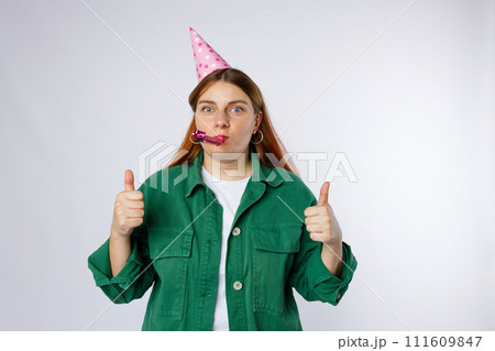 Young happy woman in birthday party hat with playing pipe showing thumbs up gesture. Emotional, young face. Human emotions, facial expression and holiday concept. Young happy woman in birthday party hat with playing pipe showing thumbs up gesture. Emotional, young face. Human emotions, facial expression and holiday concept. 111609847