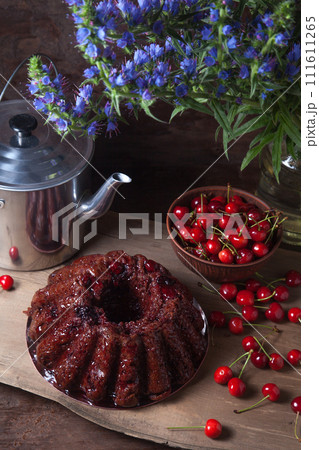 Delicious chocolate cake with sweet cherry, vintage kettle and bouquet of blue flowers on dark wooden background.. Delicious chocolate cake with sweet cherry, vintage kettle and bouquet of blue flowers on dark wooden background.. 111611265