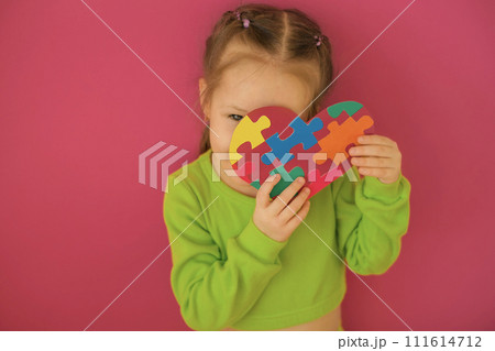 A shy sweet girl with autism spectrum disorder covers her face with a paper heart decorated with multicolored puzzles 111614712