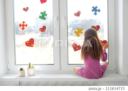 A lonely sad kid is sitting on the windowsill decorating the window of the children's room with puzzles for Autism Awareness Day A lonely sad kid is sitting on the windowsill decorating the window of the children's room with puzzles for Autism Awareness Day 111614715