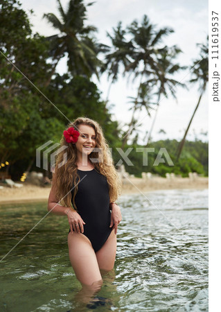Smiling woman wearing one-piece swimsuit, red flower in hair enjoys dip in ocean, tropical palms in background. Stands knee-deep in clear water, embodying beach vacation, relaxation. 111619537