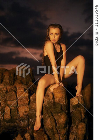 Woman in stylish black swimsuit poses on rocky shore at dusk. Model gazes into distance, ocean backdrop. Evening beachwear shoot, swim fashion. Glamor, serene escape, luxury lifestyle by sea. Woman in stylish black swimsuit poses on rocky shore at dusk. Model gazes into distance, ocean backdrop. Evening beachwear shoot, swim fashion. Glamor, serene escape, luxury lifestyle by sea. 111619546