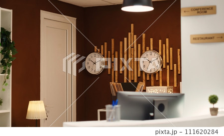 Empty cozy check in front desk in stylish luxurious resort lounge interior filled with plants and clocks on wall. Welcoming hotel reception counter ready for arriving guests Empty cozy check in front desk in stylish luxurious resort lounge interior filled with plants and clocks on wall. Welcoming hotel reception counter ready for arriving guests 111620284