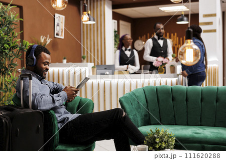 Happy smiling black guy sitting with luggage in hotel lobby in wireless headphones using digital tablet. Young African American man traveler watching movie while waiting for check-in 111620288