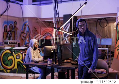 Suspicious hacker looking away after breaking into server and doing illegal activity. Young asian criminals in hood leaving abandoned warehouse after breaching database and stealing data 111620312