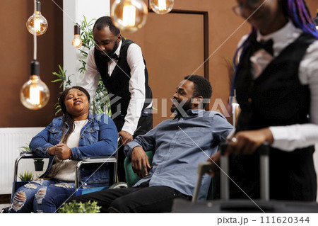 Hotel clerk talking with disabled woman in lobby, offering his assistance to guest in wheelchair. Helpful bellboy assisting tourist with disabilities during check-in or check-out 111620344
