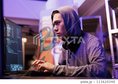 Asian criminal breaking into database and stealing confidential data. Hacker in hood using internet virus malware on computer to breach data while working in abandoned warehouse at night Asian criminal breaking into database and stealing confidential data. Hacker in hood using internet virus malware on computer to breach data while working in abandoned warehouse at night 111620348