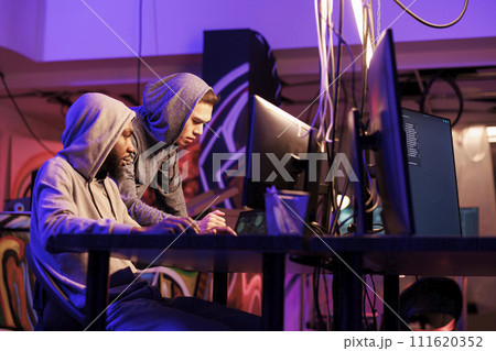 Internet scammer helping hacker to breach server and steal financial data. Two young criminals in hoods breaking law while programming illegal malicious software together Internet scammer helping hacker to breach server and steal financial data. Two young criminals in hoods breaking law while programming illegal malicious software together 111620352