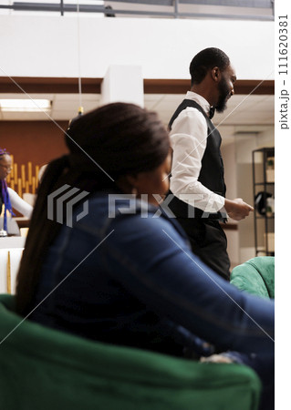 African American hotel staff and guest in lobby. Woman traveler waiting for check-in at reception area, friendly smiling bellboy helping guests with luggage. Hospitality and tourism Industry 111620381