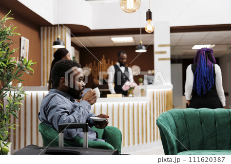 African American businessman drinking coffee while waiting for check-in procedure in hotel lobby. Tired black man traveler holding cup of espresso resting after long flight, waiting for room 111620387