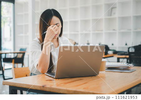 Business and technology concept, A stressed young female professional is rubbing her forehead while looking at a laptop screen in a modern workspace setting. 111621953