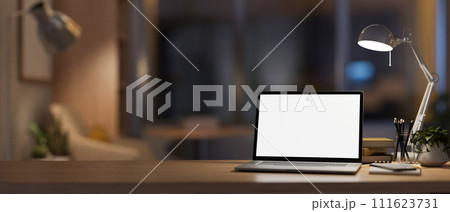 A modern, cozy home office at night with a white-screen laptop computer mockup on the table. A modern, cozy home office at night with a white-screen laptop computer mockup on the table. 111623731