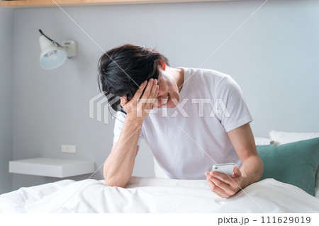 ベッドの上でスマホを見てショックを受けるアジア人男性(寝坊・遅刻・失恋・炎上・大暴落) ベッドの上でスマホを見てショックを受けるアジア人男性(寝坊・遅刻・失恋・炎上・大暴落) 111629019