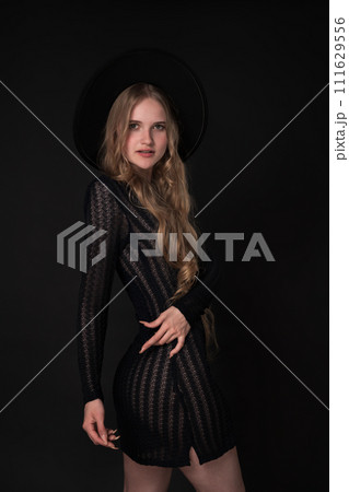 Slim woman striking graceful pose with one hand on waist. Black background accentuates her beauty, while her black short knitted translucent dress and wide brimmed felt hat add touch of elegance 111629556