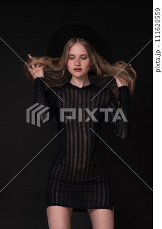 Portrait slim young blonde woman with closed eyes and hands in motion straightening long hair. Model in short tight knitted translucent dress and wide-brimmed felt hat. Black background adds to drama 111629559