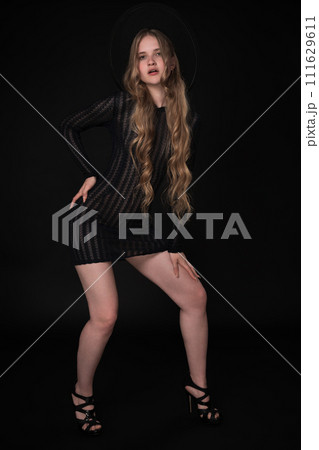 Full length of young woman with long legs, posing on black background. Blonde model with long hair is wearing black short knitted transparent tight dress, wide brimmed felt hat and high heeled shoes 111629611