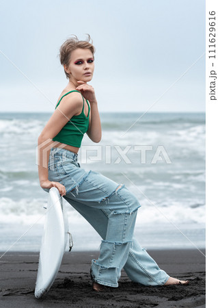 Portrait hipster woman surfer sitting on surf board lying on sand, on background of sea waves. Side view of Caucasian model with short hair and bright makeup looking at camera, wearing top and jeans 111629616