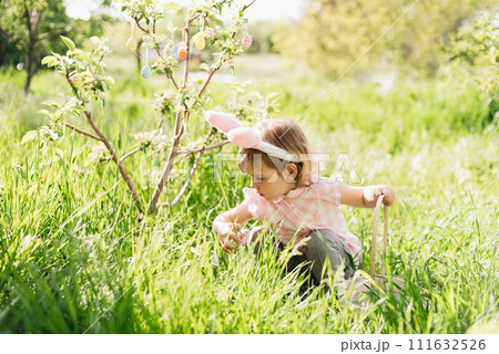 Easter egg hunt. Girl child Wearing Bunny Ears Running To Pick Up Egg In Garden. Easter tradition. Baby with basket full of colorful eggs. Easter egg hunt. Girl child Wearing Bunny Ears Running To Pick Up Egg In Garden. Easter tradition. Baby with basket full of colorful eggs. 111632526