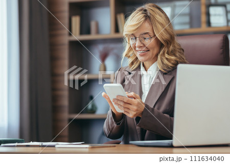 Confident mature woman in a stylish office. Smiling as she reads her smartphone screen, showcasing remote work. 111634040