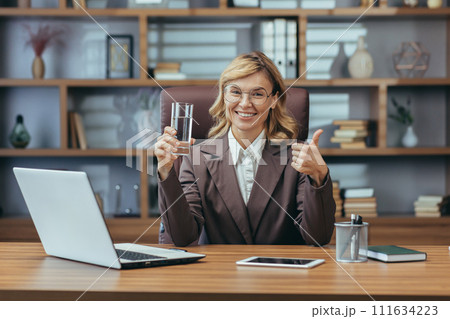 Professional senior businesswoman with glasses holding water and giving thumbs up in a modern office setting. Professional senior businesswoman with glasses holding water and giving thumbs up in a modern office setting. 111634223