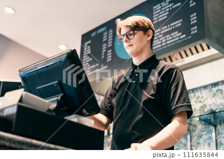Young waiter serving customer at cash point in cafe. Man working with POS terminal. Cashier, barista checking for payment receipt. Hospitality, server and preparing a slip at the till in coffee shop 111635844