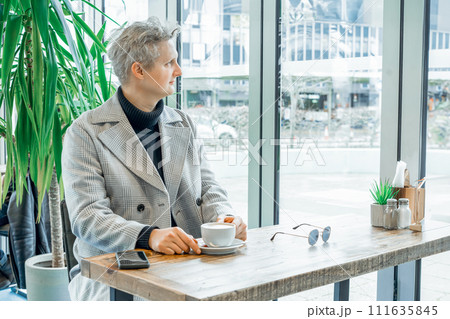 Portrait of thoughtful handsome young stylish man in checked coat relaxing with cup of coffee in modern cafe shop . Cold season elegant fashion stylish outfit trend. Retro, Old money aesthetic. 111635845