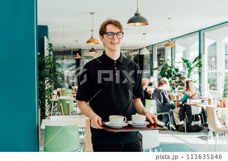 Portrait of young smiling affable waiter carrying on tray with coffee cups to client table in cafe. Hospitality service job. Occupation and work concept. Part-time work for students. Selective focus 111635846