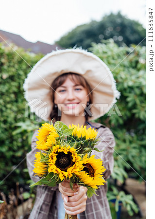 Focus on fresh sunflowers bouquet in hands of young female farmer on the green trees background. Growing flowers. Rural, Cottage core lifestyle. Vertical card. Selective focus 111635847